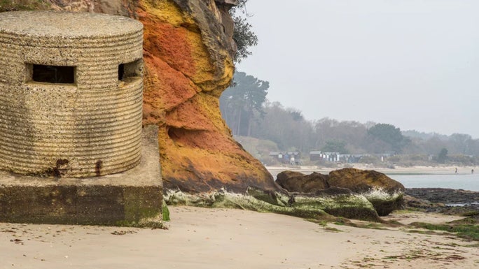 A round ribbed concrete pillbox on the shore by a golden cliff, with a line of beach huts on the curve of the bay beyond, at Studland Beach, Dorset